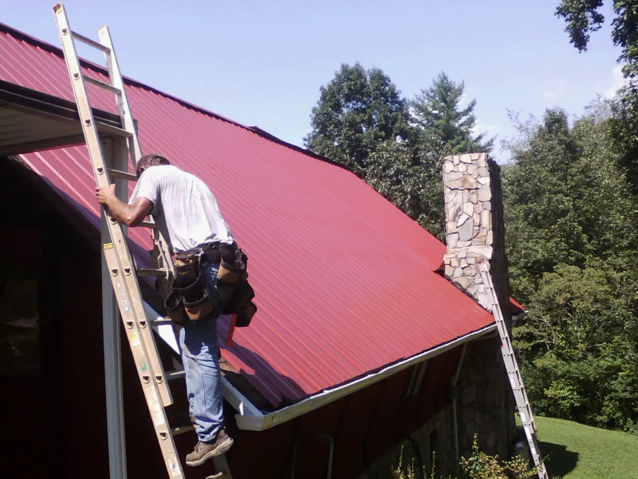 Roofer on a ladder beside a red metal roof and stone chimney — Soffit & Fascia Repair by Roofing Brothers in Garrison